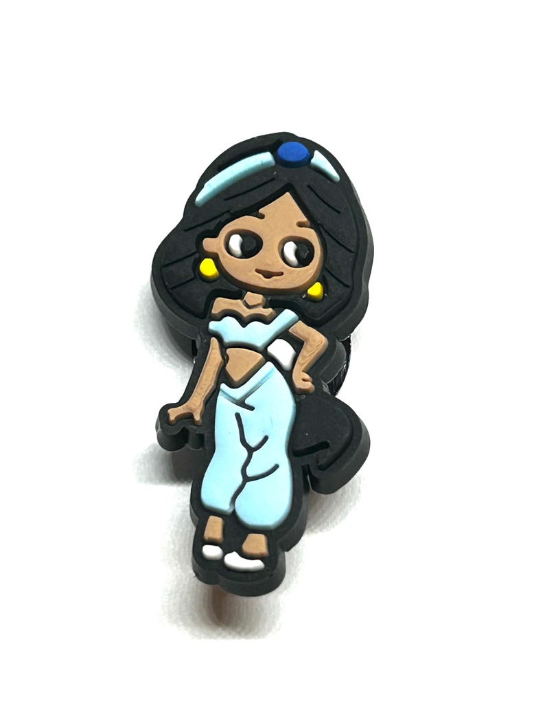 Puede incluir: Un dije de estilo caricatura que presenta un personaje con cabello negro, piel morena y ropa azul claro. El dije tiene una diadema azul con una gema azul, aretes amarillos y zapatos blancos. El personaje est&aacute; de pie.