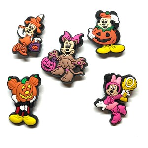 K&ouml;nnte beinhalten: Eine Sammlung von Charms mit Halloween-Motiven. Die Charms zeigen Micky und Minnie Maus und andere Charaktere in Kost&uuml;men, darunter eine Hexe, eine Katze und Charaktere, die K&uuml;rbisse halten. Die Charms sind in verschiedenen Farben erh&auml;ltlich, darunter Orange, Rosa und Braun.