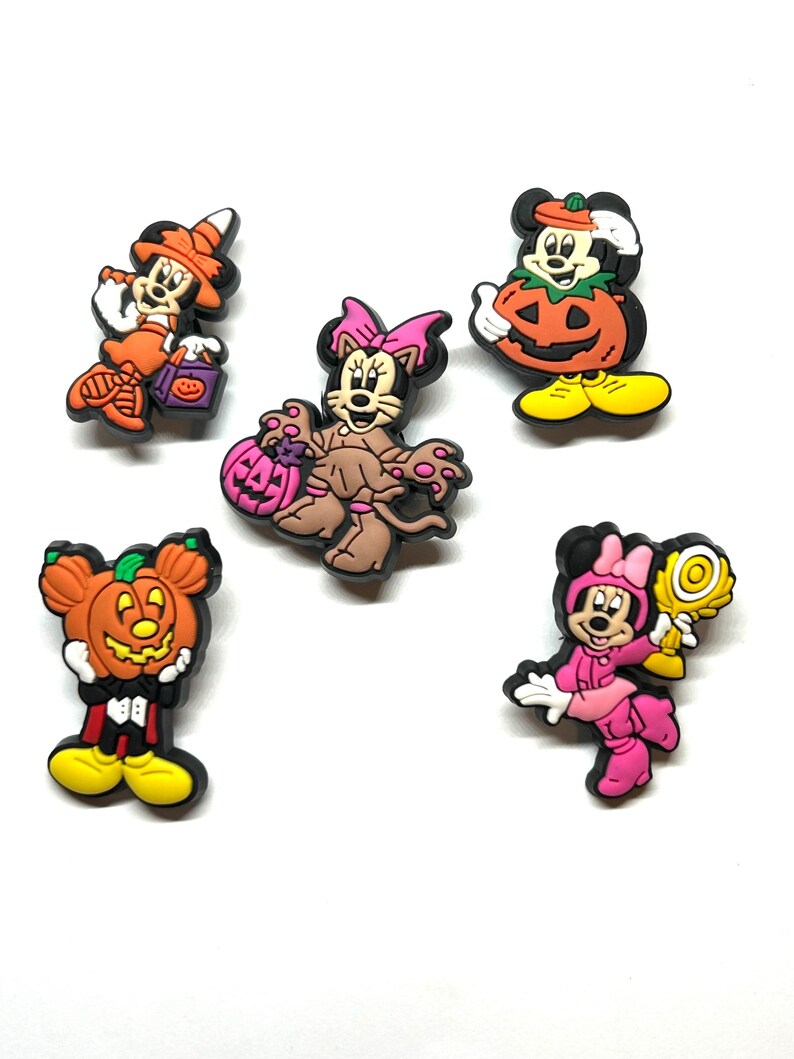 K&ouml;nnte beinhalten: Eine Sammlung von Charms mit Halloween-Motiven. Die Charms zeigen Micky und Minnie Maus und andere Charaktere in Kost&uuml;men, darunter eine Hexe, eine Katze und Charaktere, die K&uuml;rbisse halten. Die Charms sind in verschiedenen Farben erh&auml;ltlich, darunter Orange, Rosa und Braun.