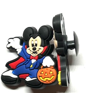 K&ouml;nnte beinhalten: Ein Halloween-Schuhanh&auml;nger mit Mickey Mouse als Vampir, der einen K&uuml;rbis h&auml;lt. Der Anh&auml;nger ist haupts&auml;chlich schwarz, mit roten, blauen, wei&szlig;en und orangefarbenen Akzenten. Zum Befestigen an Schuhen.
