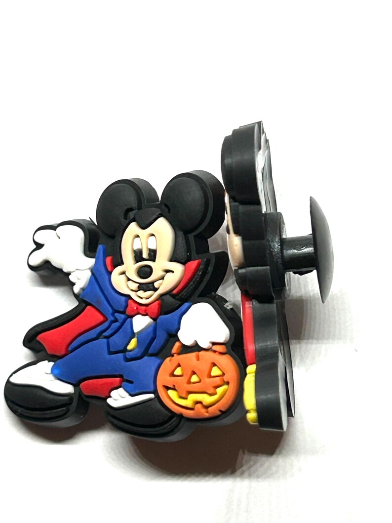K&ouml;nnte beinhalten: Ein Halloween-Schuhanh&auml;nger mit Mickey Mouse als Vampir, der einen K&uuml;rbis h&auml;lt. Der Anh&auml;nger ist haupts&auml;chlich schwarz, mit roten, blauen, wei&szlig;en und orangefarbenen Akzenten. Zum Befestigen an Schuhen.
