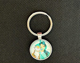 May include: A silver-colored keychain with a round pendant featuring a cartoon image of a woman in a teal dress and a tiger. The keychain has a silver ring for attaching keys.
