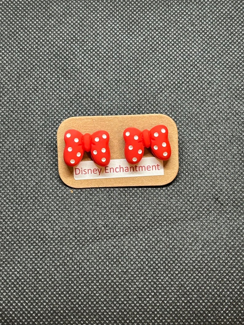 Pu&ograve; includere: Un paio di orecchini rossi a forma di fiocco con pois bianchi, presentati su un piccolo cartoncino rettangolare beige. Il cartoncino riporta la scritta "Disney Enchantment". Gli orecchini sono su uno sfondo grigio strutturato.