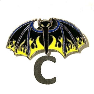 May include: A black and yellow bat-shaped enamel pin with blue accents. The pin features a stylized bat design with flame details and a silver border. A gray letter "C" is below the pin.