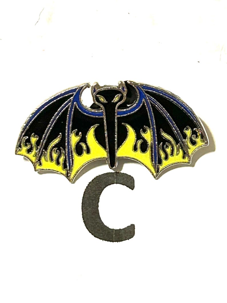 May include: A black and yellow bat-shaped enamel pin with blue accents. The pin features a stylized bat design with flame details and a silver border. A gray letter "C" is below the pin.