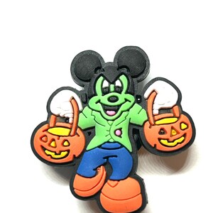 K&ouml;nnte beinhalten: Ein Halloween-Charme mit einer Cartoonfigur als Frankenstein. Die Figur ist gr&uuml;n mit schwarzen Ohren, tr&auml;gt eine gr&uuml;ne Jacke, blaue Hose und orangefarbene Schuhe. Sie h&auml;lt zwei orangefarbene K&uuml;rbis-Eimer mit wei&szlig;en Griffen.