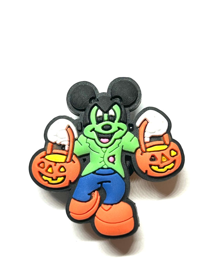 K&ouml;nnte beinhalten: Ein Halloween-Charme mit einer Cartoonfigur als Frankenstein. Die Figur ist gr&uuml;n mit schwarzen Ohren, tr&auml;gt eine gr&uuml;ne Jacke, blaue Hose und orangefarbene Schuhe. Sie h&auml;lt zwei orangefarbene K&uuml;rbis-Eimer mit wei&szlig;en Griffen.