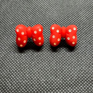 Pu&ograve; includere: Due orecchini rossi a forma di fiocco con pois bianchi. Gli orecchini sono realizzati in materiale lucido e sono su uno sfondo strutturato grigio scuro. I fiocchi sono simmetrici e hanno un design classico.