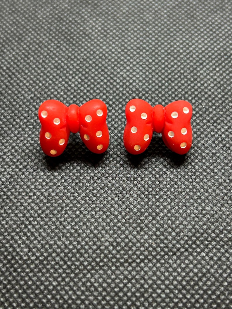 Pu&ograve; includere: Due orecchini rossi a forma di fiocco con pois bianchi. Gli orecchini sono realizzati in materiale lucido e sono su uno sfondo strutturato grigio scuro. I fiocchi sono simmetrici e hanno un design classico.