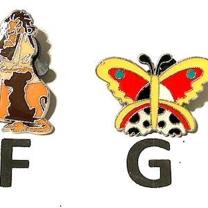 May include: Two enamel pins: one depicts a lion in shades of brown, tan, and orange, and the other a butterfly with yellow, red, black, and white details. The letters "F" and "G" are below the pins.