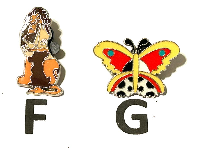 May include: Two enamel pins: one depicts a lion in shades of brown, tan, and orange, and the other a butterfly with yellow, red, black, and white details. The letters "F" and "G" are below the pins.