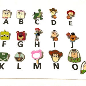 May include: A collection of colorful enamel pins featuring characters from the animated film Toy Story. The pins include characters like Woody, Buzz Lightyear, and others, each with a unique design and vibrant colors. The pins are arranged on a white surface.