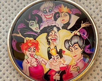 May include: A round silver keychain pendant featuring a colorful illustration of several cartoon villains. The characters include a woman with a crown, a woman with a purple octopus, and a woman with a black and white hairstyle.