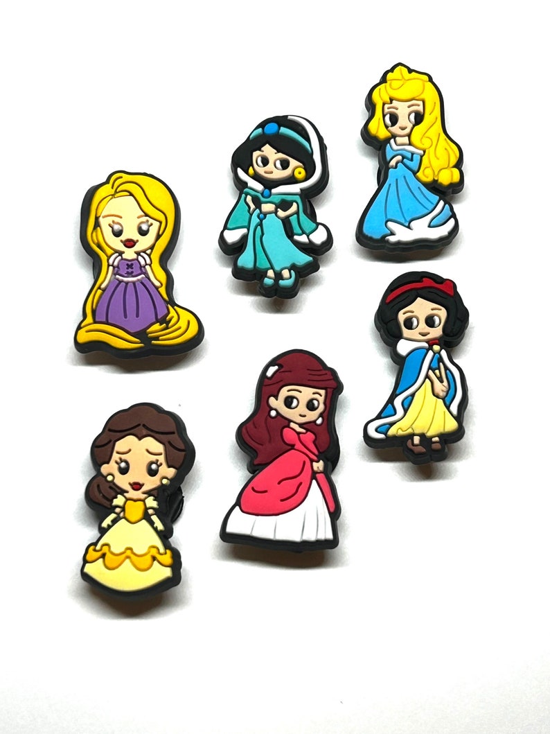 Puede incluir: Una colecci&oacute;n de coloridos dijes de zapatos de personajes de dibujos animados. Los dijes presentan a varias princesas con sus atuendos caracter&iacute;sticos, incluyendo a Rapunzel, Jasmine, Aurora, Blancanieves, Bella y otras. Cada dije tiene un borde negro.