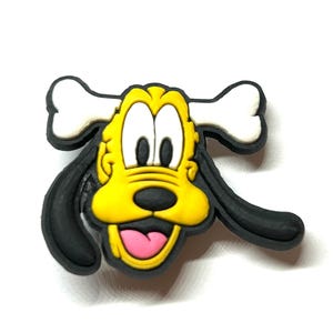 K&ouml;nnte beinhalten: Ein farbenfrohes Cartoon-Hunde-Gesicht mit einem Knochen oben. Der Hund hat ein gelbes Gesicht, schwarze Ohren und eine rosa Zunge. Der Knochen ist wei&szlig;. Das Design ist wahrscheinlich f&uuml;r einen Schuhanh&auml;nger oder ein &auml;hnliches Accessoire.