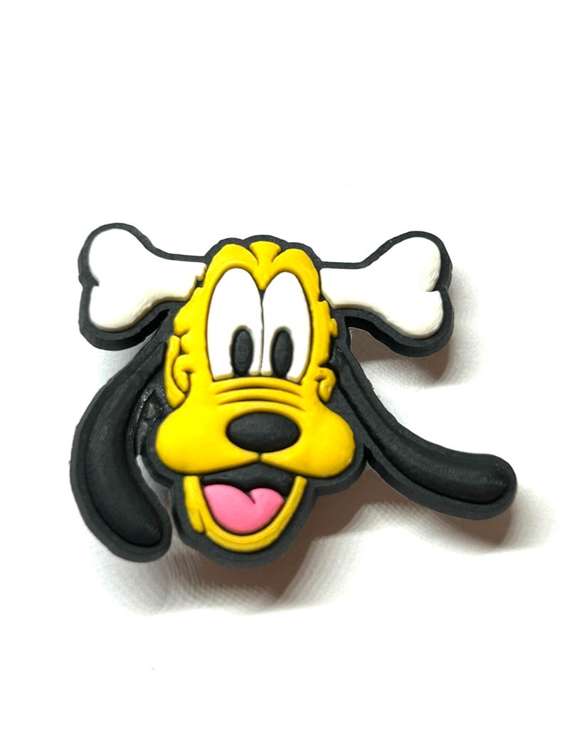 K&ouml;nnte beinhalten: Ein farbenfrohes Cartoon-Hunde-Gesicht mit einem Knochen oben. Der Hund hat ein gelbes Gesicht, schwarze Ohren und eine rosa Zunge. Der Knochen ist wei&szlig;. Das Design ist wahrscheinlich f&uuml;r einen Schuhanh&auml;nger oder ein &auml;hnliches Accessoire.