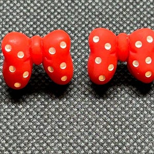 Pu&ograve; includere: Due accessori rossi a forma di fiocco con pois bianchi. I fiocchi sono realizzati in un materiale liscio e sono posizionati su una superficie strutturata grigio scuro. Potrebbero essere usati come abbellimenti per l'artigianato o gli accessori.