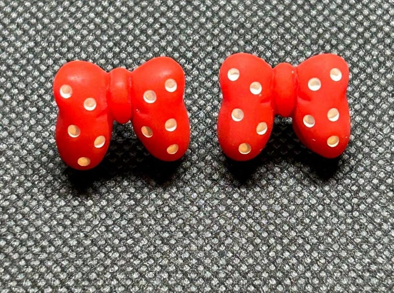 Pu&ograve; includere: Due accessori rossi a forma di fiocco con pois bianchi. I fiocchi sono realizzati in un materiale liscio e sono posizionati su una superficie strutturata grigio scuro. Potrebbero essere usati come abbellimenti per l'artigianato o gli accessori.