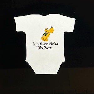 Könnte beinhalten: Weißer Baby-Body mit einem Cartoon-Bild eines gelben Hundes mit grünem Rucksack. Der Text "It's Ruff Being This Cute" ist darunter in schwarzer Schrift gedruckt. Der Body hat kurze Ärmel und einen runden Ausschnitt. Babykleidung.