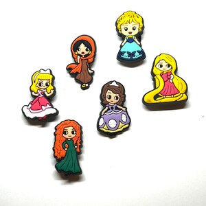 Puede incluir: Una colecci&oacute;n de amuletos de personajes de dibujos animados coloridos, con varias princesas con diferentes atuendos. Los amuletos est&aacute;n hechos de un material brillante y est&aacute;n sobre un fondo blanco. Cada amuleto tiene un dise&ntilde;o y una combinaci&oacute;n de colores &uacute;nicos.