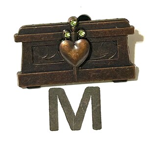 May include: A small, bronze-colored decorative chest with a heart-shaped clasp and three green gemstones. The chest has a rectangular shape with ornate detailing and is accompanied by a gray letter "M".