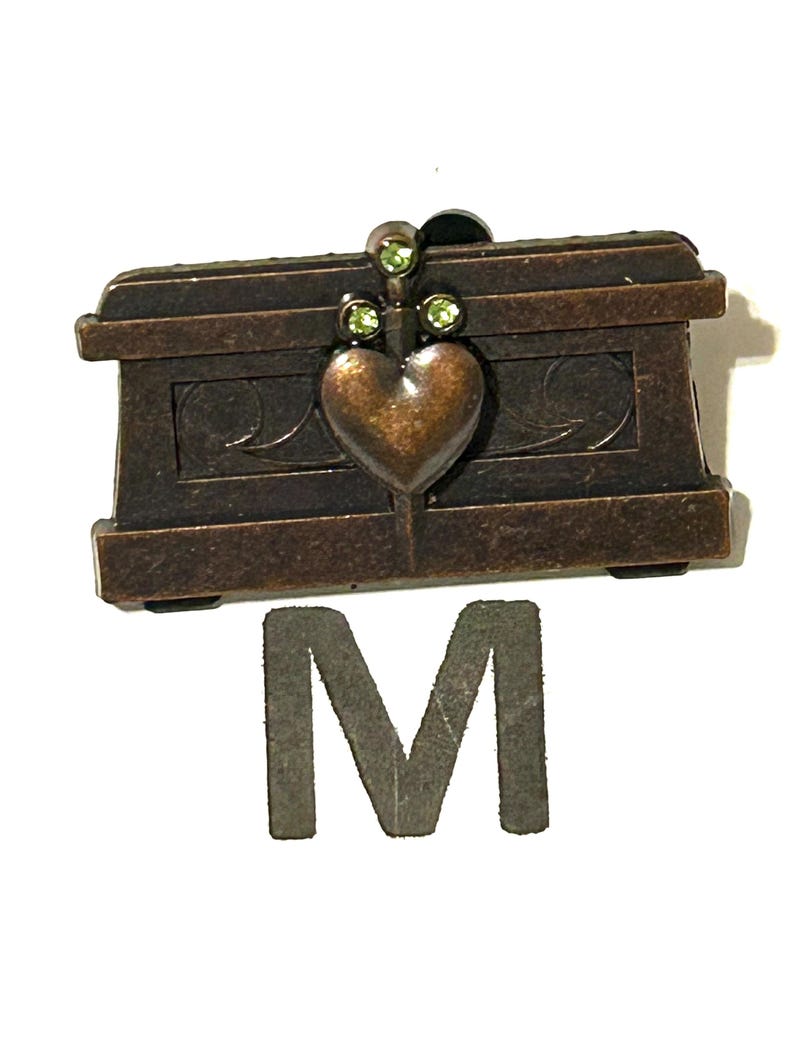 May include: A small, bronze-colored decorative chest with a heart-shaped clasp and three green gemstones. The chest has a rectangular shape with ornate detailing and is accompanied by a gray letter "M".