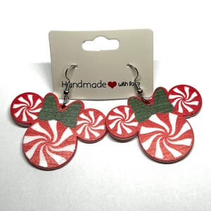 Op de afbeelding: Een paar feestelijke oorbellen in de vorm van Mickey Mouse-hoofden, met rood-witte pepermunt snoepontwerpen. De oorbellen hebben groene strikaccenten en zilveren haken. Het label luidt "Handmade with love".