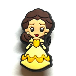Puede incluir: Un dije de dibujos animados que representa a una princesa con un vestido amarillo y un contorno negro. La princesa tiene el pelo casta&ntilde;o y pendientes dorados. El dije probablemente est&aacute; hecho de goma o un material similar.
