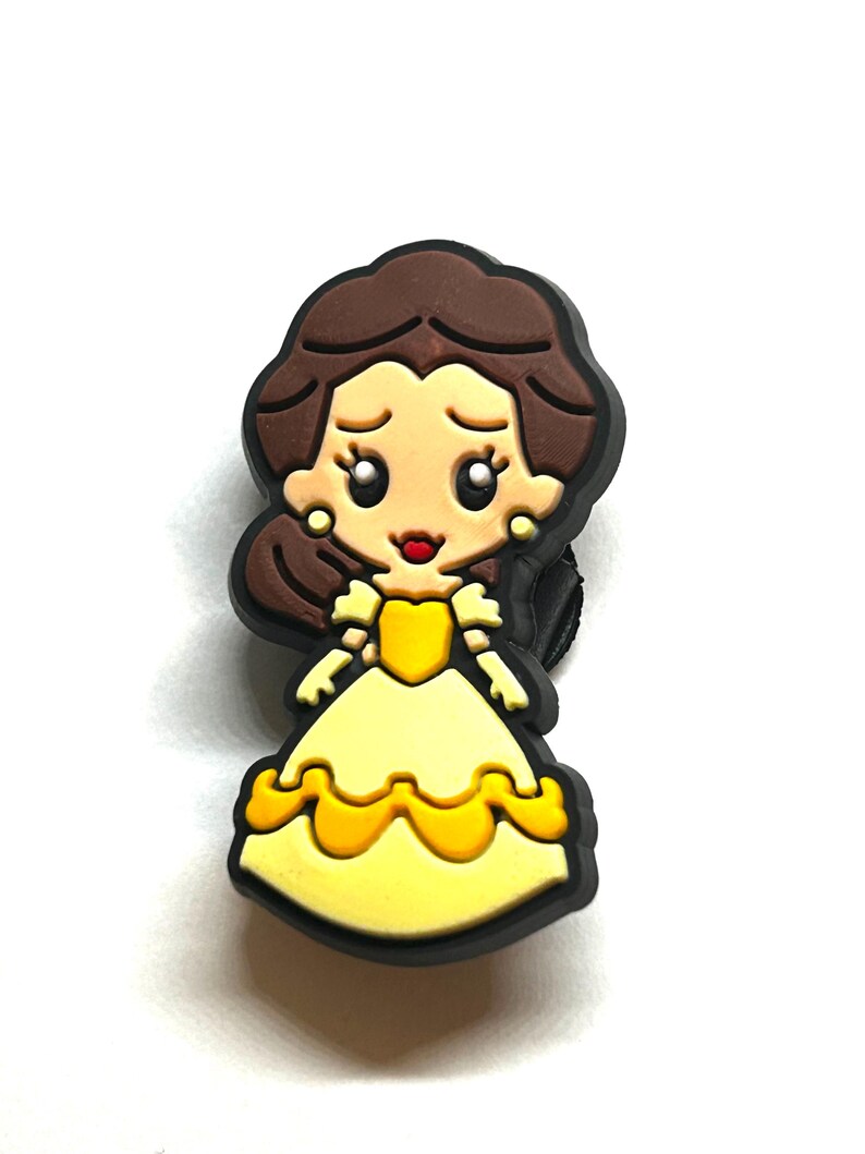 Puede incluir: Un dije de dibujos animados que representa a una princesa con un vestido amarillo y un contorno negro. La princesa tiene el pelo casta&ntilde;o y pendientes dorados. El dije probablemente est&aacute; hecho de goma o un material similar.