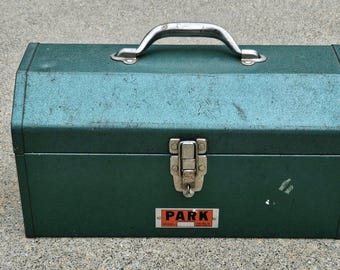 May include: A vintage green metal toolbox with a silver handle and latch. The toolbox has a rectangular shape with a slightly curved top. A small rectangular label with the word "PARK" is on the front.