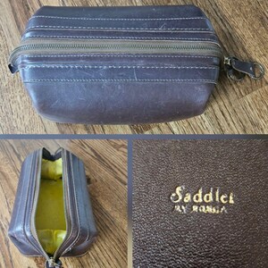 May include: A brown leather travel case with a zipper closure and a small handle. The case is rectangular with a rounded top and features white stitching details. The interior is lined with yellow fabric. The text "Saddler by Roma" is embossed on the leather.