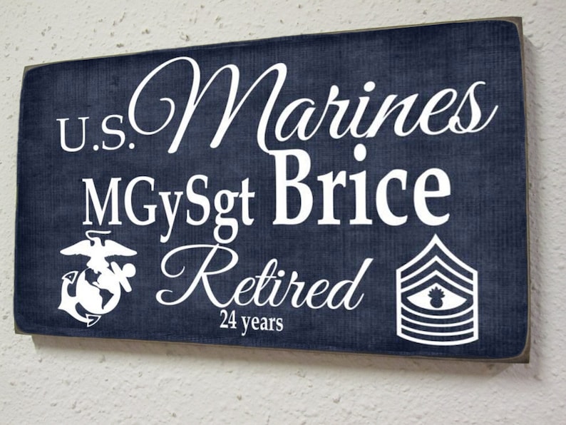 Puede incluir: Un letrero de madera azul marino con texto blanco que dice "U.S. Marines MGySgt Brice Retired 24 years". El letrero tambi&eacute;n presenta un emblema blanco del Cuerpo de Marines y un gal&oacute;n blanco con una pi&ntilde;a.
