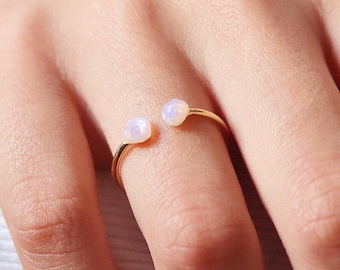 May include: A delicate gold ring featuring two small, round, opalescent gemstones. The ring's band is thin and curves around the finger, with the gemstones positioned on either side of the finger. The gemstones have a soft, milky white color.