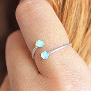 Könnte beinhalten: Ein silberfarbener Ring mit zwei hellblauen, runden Edelsteinen. Der Ring umschließt den Finger, wobei sich die Edelsteine auf beiden Seiten befinden. Der Ring hat ein minimalistisches Design.