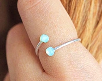 May include: A silver-colored ring with two light blue, round gemstones. The ring wraps around the finger with the gemstones positioned on either side. The ring is a minimalist design.