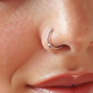 Peut inclure: Un anneau de nez circulaire de couleur argentée doté d'une petite fermeture à bille lisse. Le bijou est placé dans la narine, formant une boucle continue avec une finition métallique polie qui reflète la lumière sur la surface de la peau.
