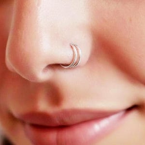 May include: A double hoop nose ring made of thin, polished silver-toned metal. The jewelry consists of two separate, circular bands that sit closely together, creating a layered appearance when worn in the nostril piercing of the nose.