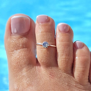 Puede incluir: Un anillo de plata para el dedo del pie con diseño de concha. El anillo se lleva en un pie, sobre un fondo azul. El anillo es pequeño y delicado, con un dije de concha detallado. El pie tiene un tono de piel claro y está mojado.