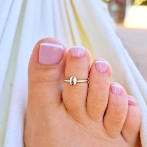 Puede incluir: Un anillo de plata para el dedo gordo del pie con un pequeño diseño de concha ovalada. El anillo se lleva en un pie con esmalte de uñas rosa y un acento blanco en las uñas de los pies. El fondo es una tela blanca suave.