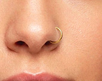 May include: A thin, circular gold nose ring hoop is inserted into the nostril. The jewelry features a smooth, polished metallic finish and a simple, continuous band design that rests securely against the skin of the nose.