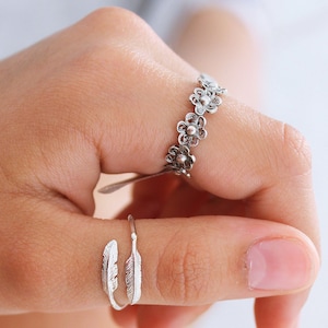 Peut inclure: Deux bagues en argent sur un doigt. L'une est une bande délicate avec une rangée de petits motifs floraux. L'autre est une bande simple avec un motif de plume.