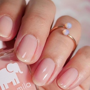 Può includere: Primo piano di una mano con smalto per unghie rosa chiaro. Le unghie sono curate e hanno una finitura lucida. La mano indossa un delicato anello d'oro con due piccole pietre di opale.