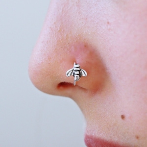 May include: A silver nose ring with a bee design. The bee is facing forward with its wings spread out.