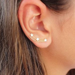 Puede incluir: Pendientes trepadores dorados con tres estrellas blancas. Los pendientes se curvan a lo largo de la oreja, creando un diseño celestial. Las estrellas están espaciadas uniformemente, añadiendo un toque de fantasía a la joya.