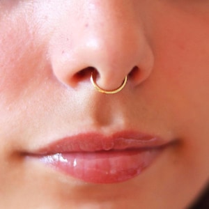 Pode incluir: Um close-up do rosto de uma pessoa com um piercing de septo dourado. O piercing tem um design simples e curvo. Os lábios da pessoa são visíveis, com um batom brilhante, vermelho-rosado. O tom de pele é claro.