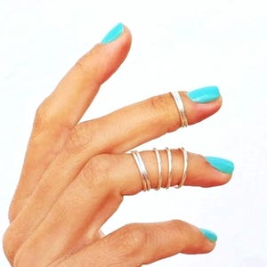 May include: A hand with turquoise fingernails displays several silver rings. One finger has a single ring, while another has a set of five thin rings. The rings are simple and minimalist in design.
