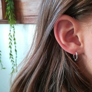 May include: Silver hoop earrings with a textured design. The earrings are worn in the earlobe.