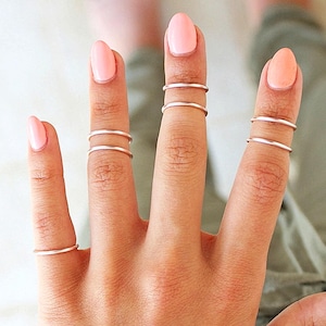 May include: A hand with several fingers adorned with delicate, thin silver rings. The rings are worn as midi rings, adding a minimalist and stylish touch. The nails are painted a soft pink, complementing the silver jewelry.