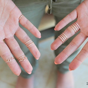 May include: A hand model wearing multiple thin gold wire rings on several fingers. The rings are simple and delicate, with some featuring a curved or looped design.