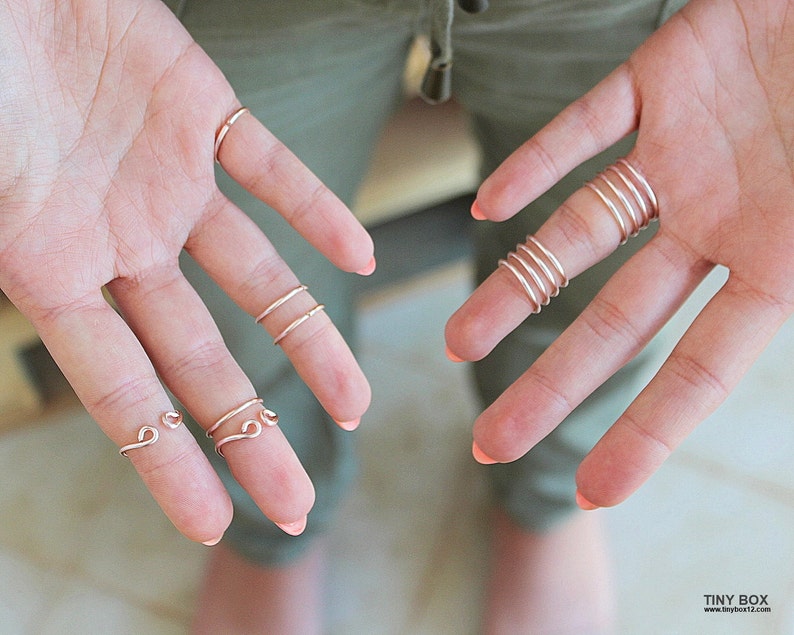 May include: A hand model wearing multiple thin gold wire rings on several fingers. The rings are simple and delicate, with some featuring a curved or looped design.